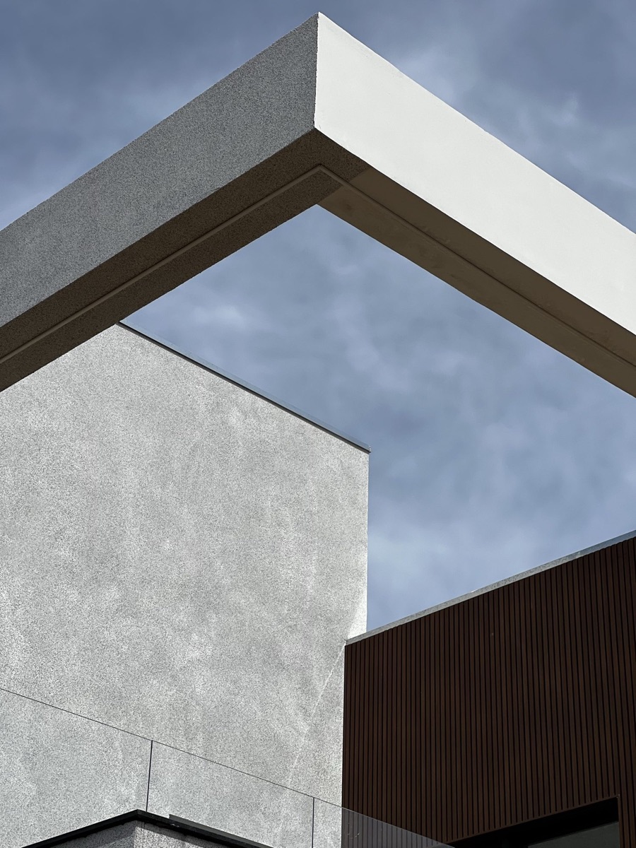 Detalhe de fachada com pórtico branco, parede em textura mineral e painel de madeira ripada contra o céu