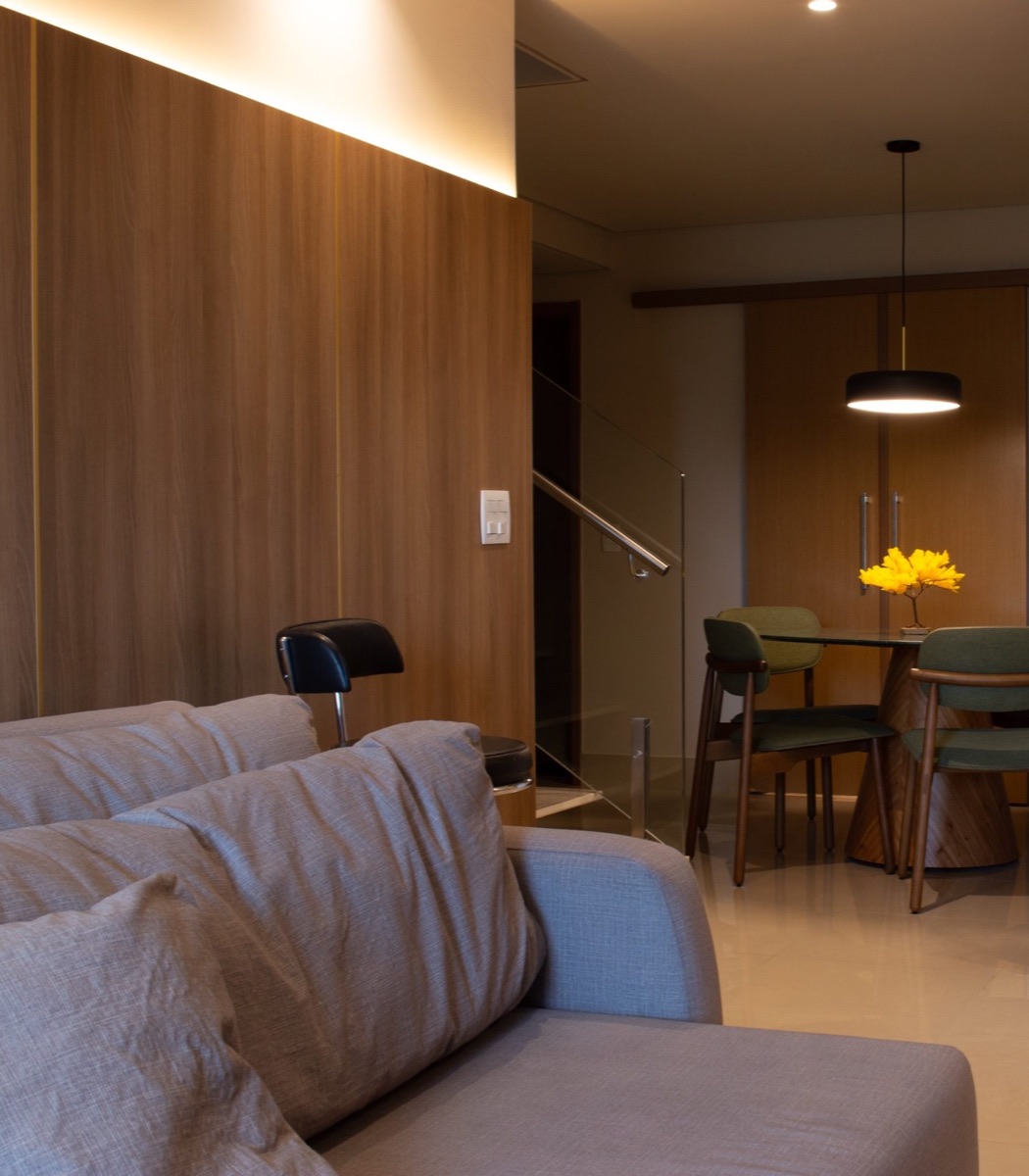 Sala de estar com painel de madeira e luz embutida, sofá cinza e mesa de jantar ao fundo
